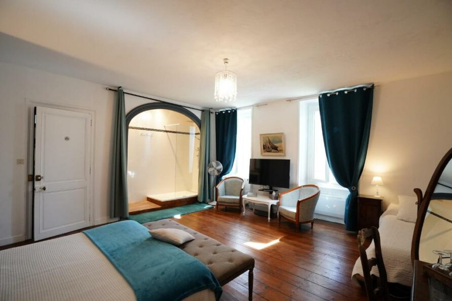 Vue de la chambre depuis un coin de la pièce. Face aux fenêtres fermées et rideaux ouverts. La salle de bain donne sur la pièce.